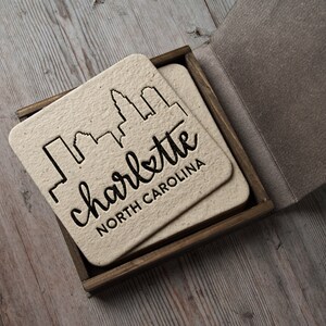 May include: Set of four coasters in a wooden box. Each coaster features a black silhouette of the Charlotte, North Carolina skyline with the text "charlotte NORTH CAROLINA".