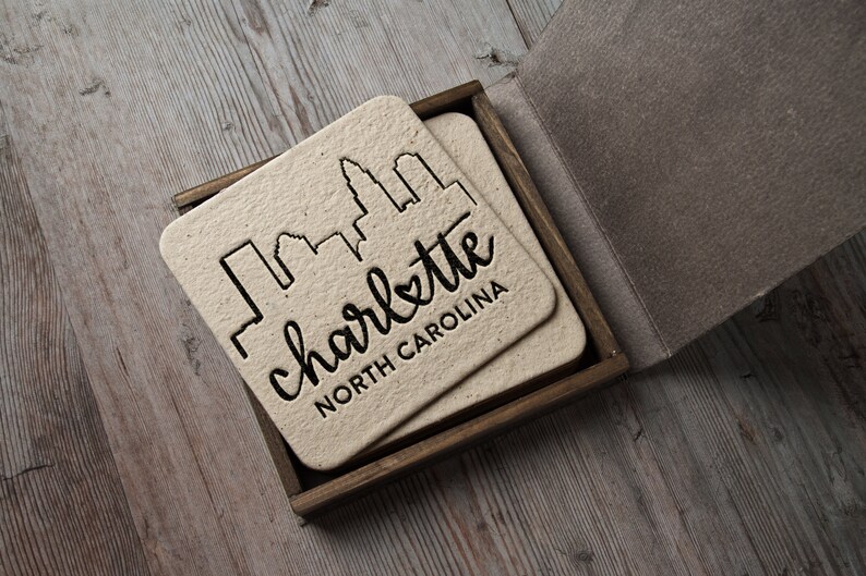 May include: Set of four coasters in a wooden box. Each coaster features a black silhouette of the Charlotte, North Carolina skyline with the text "charlotte NORTH CAROLINA".
