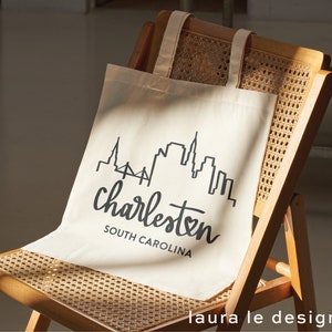 May include: A white canvas tote bag with black text and a skyline graphic. The text reads "Charleston, South Carolina".