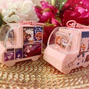 May include: Two pink miniature food trucks with clear tops. The trucks feature anime-style illustrations and text, including "GD x Gude" and "Crumbs and Kisses." Each truck has a pink keychain attached. The background includes pink flowers.