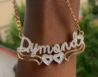 May include: Gold chain necklace with a nameplate pendant that says "Dymon" with two small hearts on the bottom. The nameplate is set with clear cubic zirconia stones.