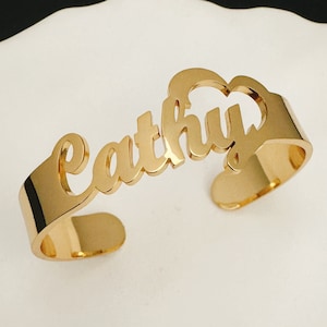 May include: Gold-toned metal bracelet with the name "Cathy" cut out in the metal. The bracelet is open and curved to fit a wrist.