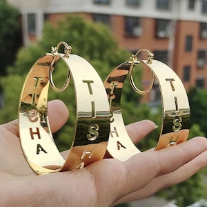 Puede incluir: Pendientes de aro dorados con la palabra "THIS IS HA" recortada en el metal.