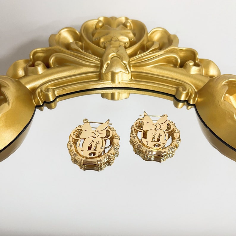 K&ouml;nnte beinhalten: Ein Paar goldfarbene Creolen mit einem Mickey-Maus-Silhouetten-Design. Die Ohrringe sind rund und haben eine strukturierte Oberfl&auml;che.