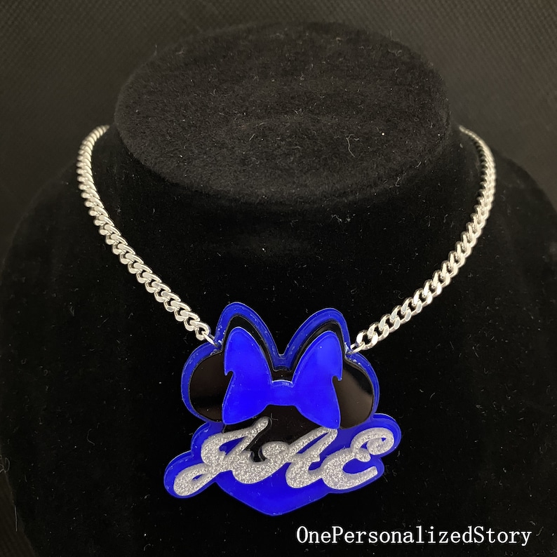 Op de afbeelding: Een zilveren ketting met een blauwe en zwarte Minnie Mouse-hanger. De hanger heeft de naam "Jace" in zilveren glitter erop.