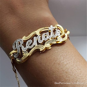 May include: A gold and silver nameplate bracelet with the name "Renate" in a decorative font. The bracelet is on a gold chain and is worn on a wrist. The nameplate has a gold border with a silver name.