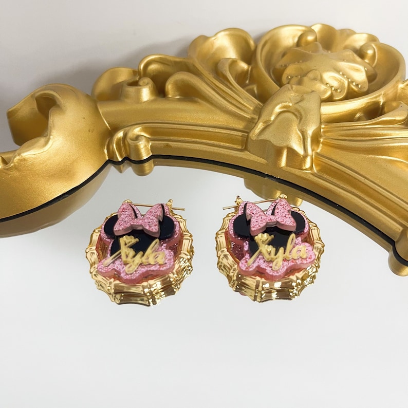 Puede incluir: Aretes de aro dorados con un dise&ntilde;o de Minnie Mouse rosa y negro y el nombre "Kyla" en letras doradas.