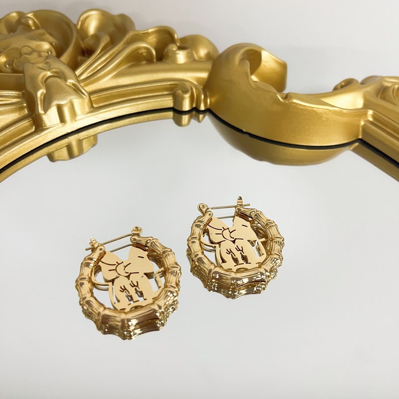 K&ouml;nnte beinhalten: Ein Paar goldene Creolen mit einem detaillierten Design eines stilisierten Elefanten in einem Kreis. Die Ohrringe sind aus Metall und haben eine gl&auml;nzende Oberfl&auml;che.