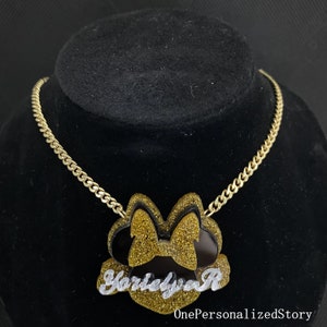 Op de afbeelding: Gouden ketting met een zwarte en gouden glitter Minnie Mouse hanger met de naam "Harleyva R" in witte cursieve letters.