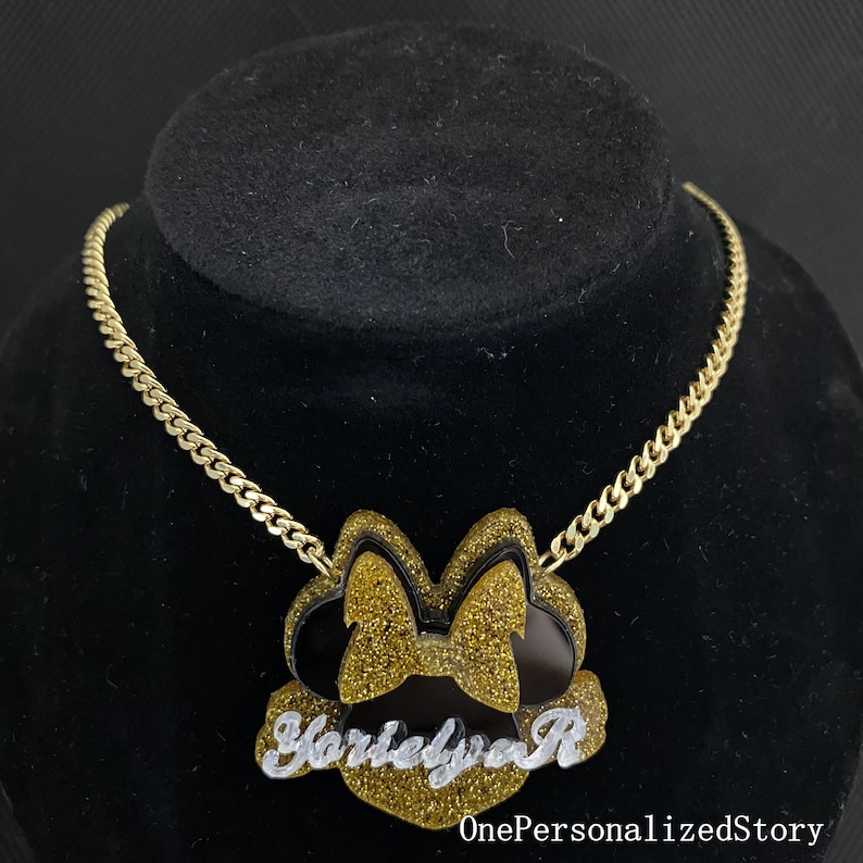 Op de afbeelding: Gouden ketting met een zwarte en gouden glitter Minnie Mouse hanger met de naam "Harleyva R" in witte cursieve letters.