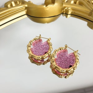Puede incluir: Un par de pendientes de aro dorados con un dise&ntilde;o de flor de purpurina rosa. Los pendientes est&aacute;n hechos de un material similar al bamb&uacute;.