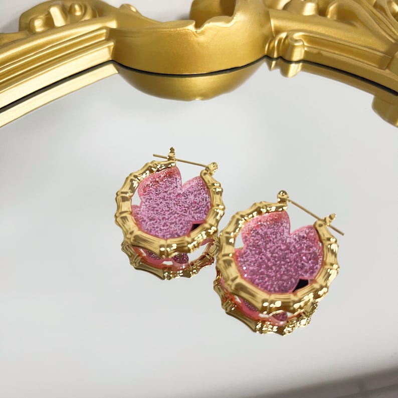 Puede incluir: Un par de pendientes de aro dorados con un dise&ntilde;o de flor de purpurina rosa. Los pendientes est&aacute;n hechos de un material similar al bamb&uacute;.