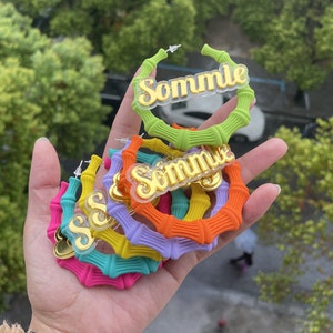 Può includere: Un set di orecchini a cerchio in bambù colorati con la parola "Sommie" scritta in lettere dorate su ogni cerchio.
