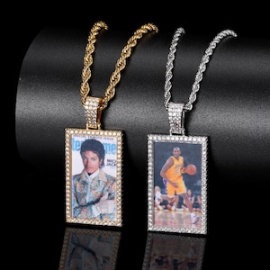 Può includere: Due collane con ciondoli. Una presenta una catena dorata e un ciondolo rettangolare con una foto di Michael Jackson. L'altra ha una catena argentata e un ciondolo con una foto di Kobe Bryant. Entrambi i ciondoli sono incastonati con piccole pietre trasparenti.