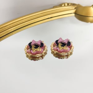 Puede incluir: Un par de pendientes de tono dorado con un dise&ntilde;o de Minnie Mouse rosa y negro. Los pendientes tienen un lazo rosa, orejas negras y la palabra "Bulla" en letras doradas.