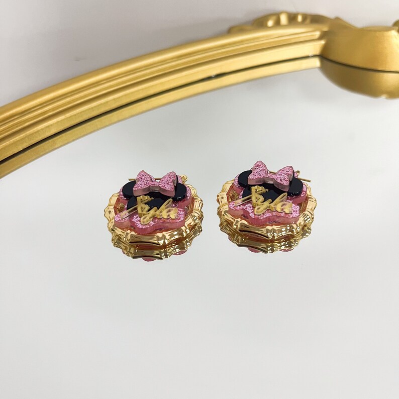 Puede incluir: Un par de pendientes de tono dorado con un dise&ntilde;o de Minnie Mouse rosa y negro. Los pendientes tienen un lazo rosa, orejas negras y la palabra "Bulla" en letras doradas.