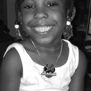 Op de afbeelding: Een zwart-witfoto van een jong meisje met een brede glimlach. Ze draagt een wit tanktop met een zilveren ketting met de tekst "Madison". Haar haar zit in vlechten en ze draagt oorbellen.