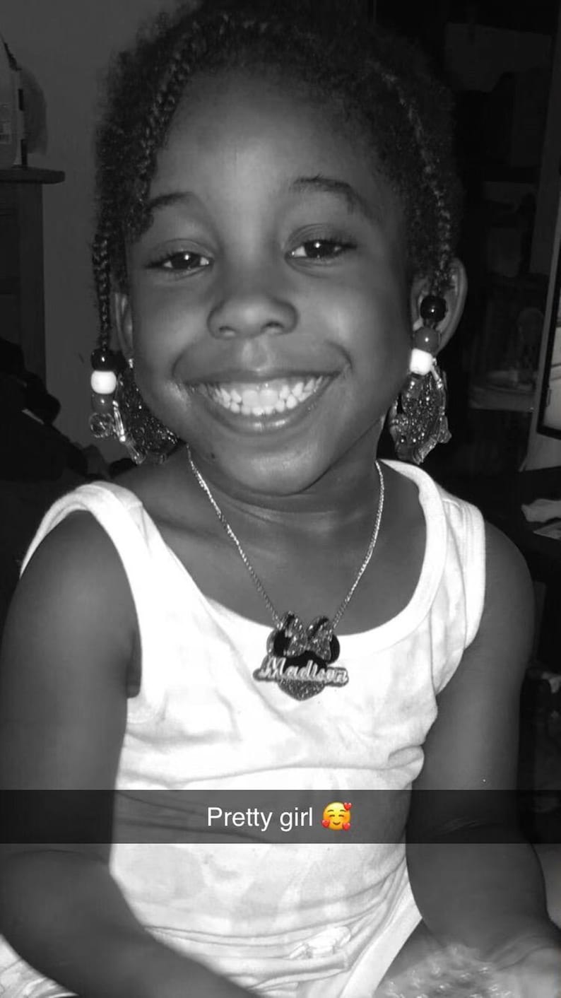 Op de afbeelding: Een zwart-witfoto van een jong meisje met een brede glimlach. Ze draagt een wit tanktop met een zilveren ketting met de tekst "Madison". Haar haar zit in vlechten en ze draagt oorbellen.