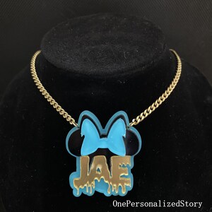 Op de afbeelding: Gouden ketting met een blauwe en zwarte hanger met een Mickey Mouse-silhouet met een strik en de naam "JAE" in gouden, druipende letters.