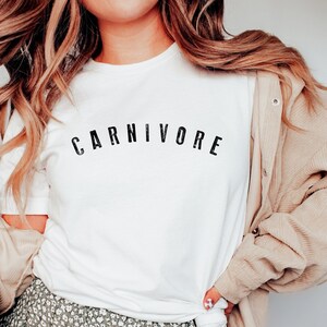 May include: A white t-shirt with the word "CARNIVORE" printed in black letters across the chest. The shirt is being worn by a person with long blonde hair and a floral skirt.