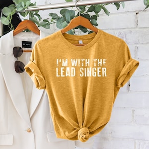 May include: A mustard yellow t-shirt with the text "I'm with the Lead Singer" printed in white on the front.