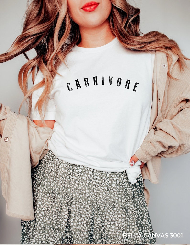 May include: A white t-shirt with the word "CARNIVORE" printed in black letters across the chest. The shirt is being worn by a person with long blonde hair and a floral skirt.