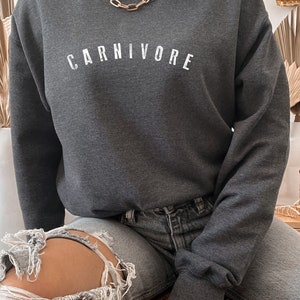 May include: A dark heather gray sweatshirt with the word "CARNIVORE" printed in white on the front. The sweatshirt is made by Gildan and is a size 18000.