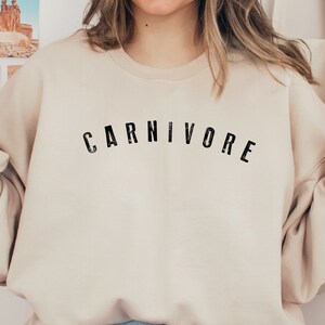 May include: A beige sweatshirt with the word "CARNIVORE" printed in black on the front. The sweatshirt is made by Gildan and is in the color "18000 Sand".