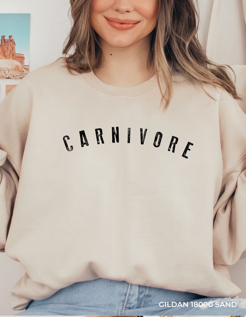 May include: A beige sweatshirt with the word "CARNIVORE" printed in black on the front. The sweatshirt is made by Gildan and is in the color "18000 Sand".