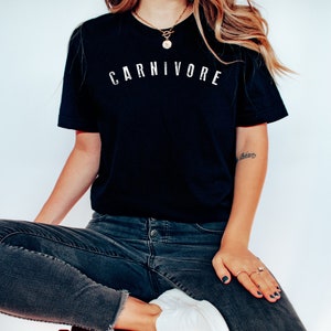 May include: A black t-shirt with the word "CARNIVORE" printed in white on the front. The shirt is being worn by a person sitting on a wooden stool.