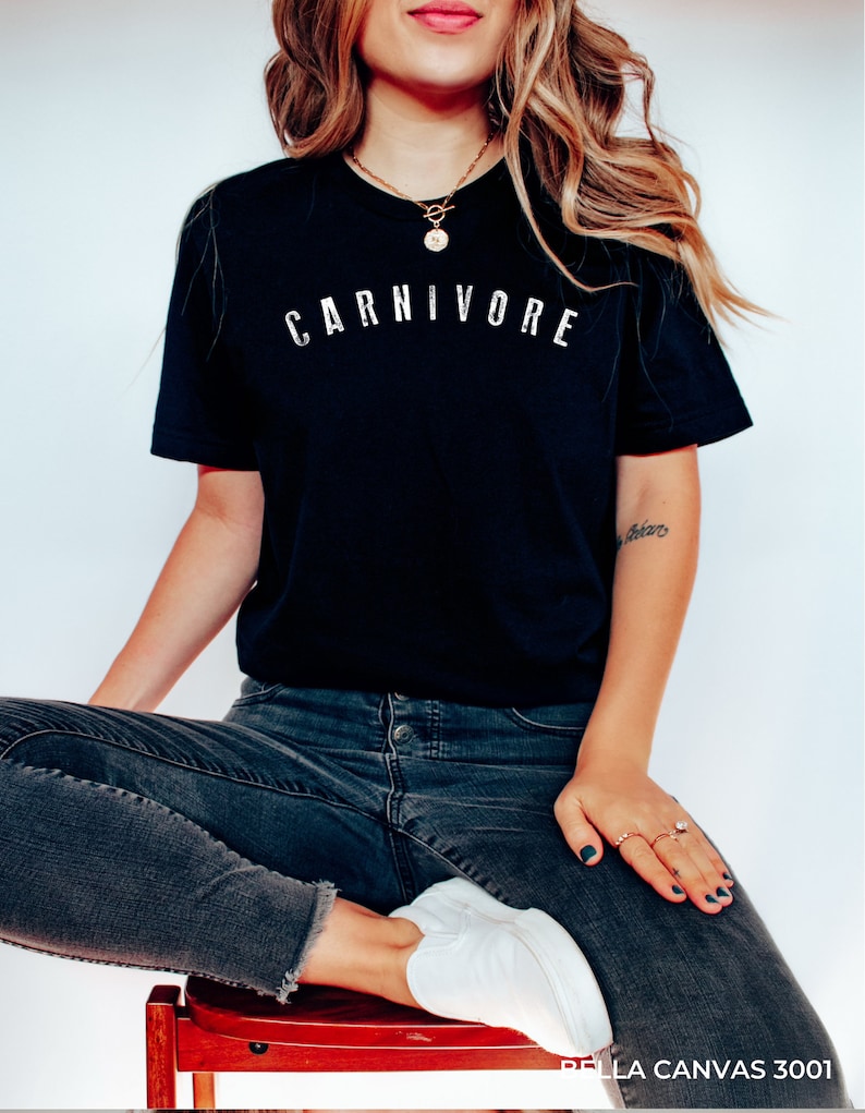 May include: A black t-shirt with the word "CARNIVORE" printed in white on the front. The shirt is being worn by a person sitting on a wooden stool.