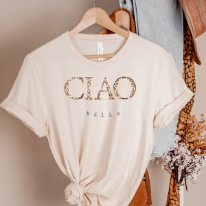 Puede incluir: Una camiseta beige con la palabra "CIAO" en letras de estampado de leopardo y la palabra "BELLA" debajo. La camiseta est&aacute; colgada de una percha de madera.