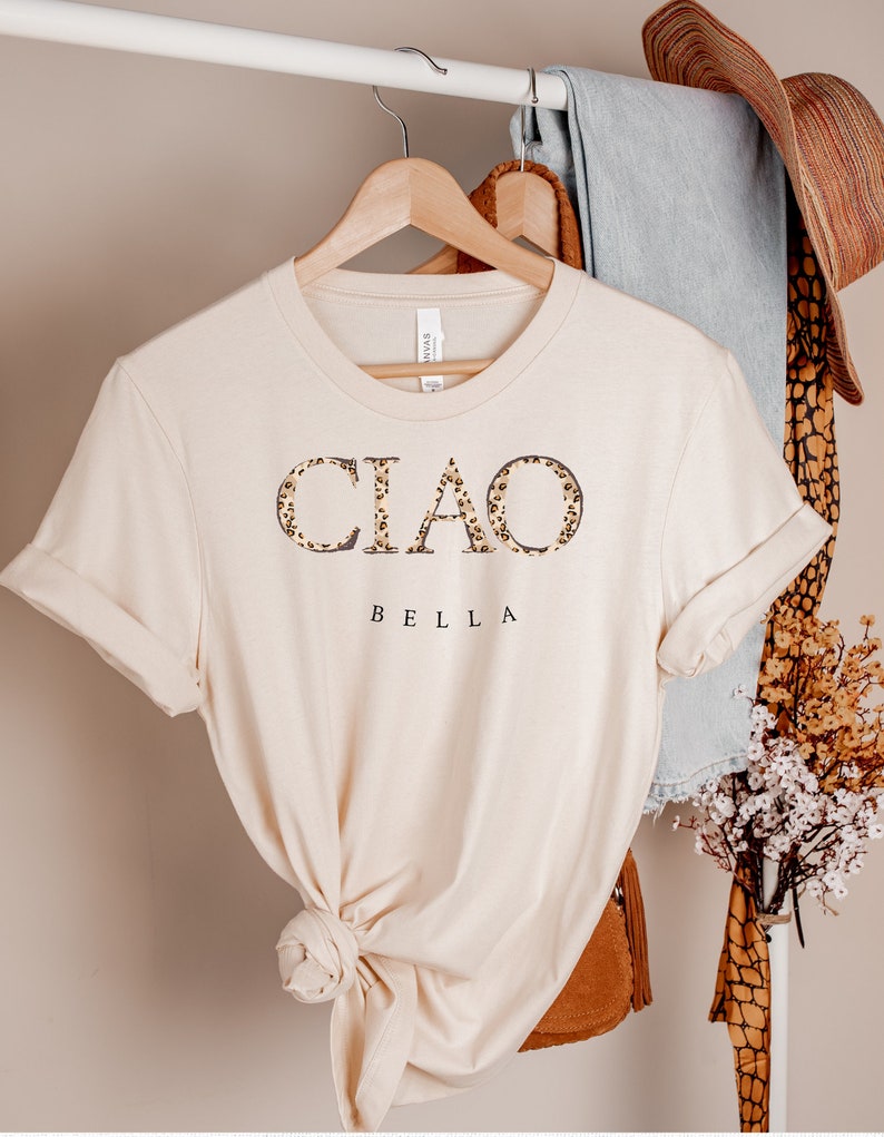 Puede incluir: Una camiseta beige con la palabra "CIAO" en letras de estampado de leopardo y la palabra "BELLA" debajo. La camiseta est&aacute; colgada de una percha de madera.