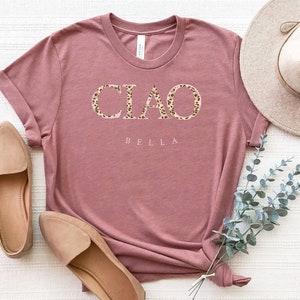 Puede incluir: Una camiseta color malva con el texto "CIAO BELLA" en letras de estampado de leopardo.
