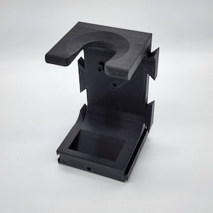 May include: Black plastic 3D printed phone stand with a curved top and a rectangular base. The stand has a small hole on the back.