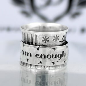 May include: A silver spinning ring with the words "I am enough" engraved on it. The ring is decorated with a snowflake design.