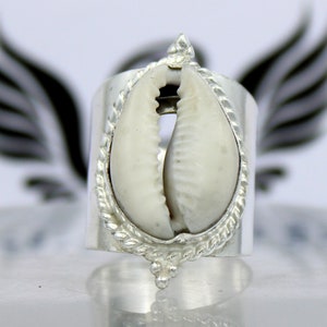 Peut inclure: Une bague en argent avec un coquillage de cauris blanc au centre. La bague est décorée de détails argentés complexes autour du coquillage.
