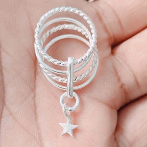 May include: A silver ring set featuring four stacked bands, three with a twisted rope design and one smooth. A small star charm dangles from the ring. The ring is shown against a light background.