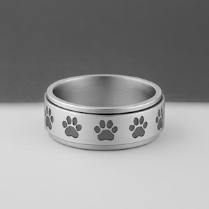 May include: A silver-colored ring with a band featuring a repeating pattern of paw prints. The ring has a smooth, polished finish and a rotating center band. The paw prints are dark gray and evenly spaced around the ring.