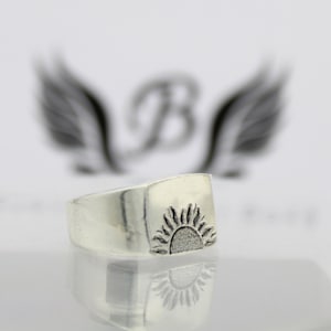 May include: A silver ring with a raised sun design. The ring is square shaped and has a smooth, polished finish.