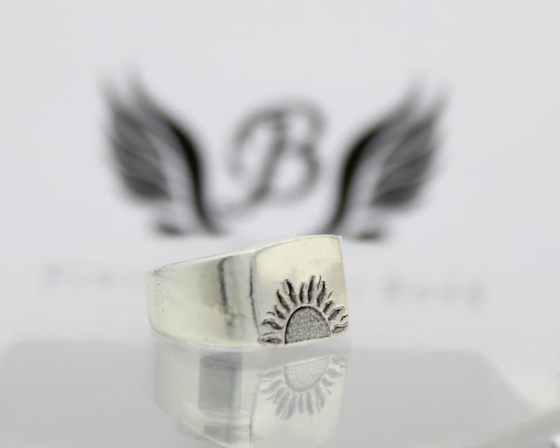 May include: A silver ring with a raised sun design. The ring is square shaped and has a smooth, polished finish.
