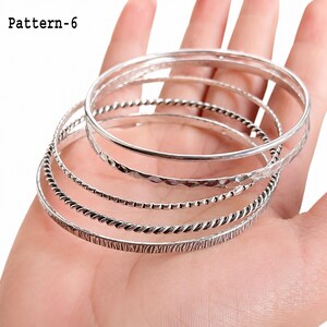 Handmade silver bangles arranged together in the palm, showing the polished edges, engraved patterns, and slim profiles.