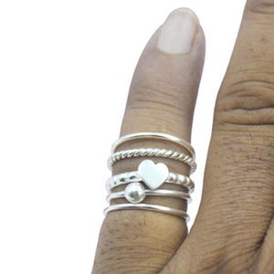 May include: A set of five silver rings stacked on a finger. The rings are different styles, including a plain band, a rope design, a beaded ring, and a ring with a heart charm.
