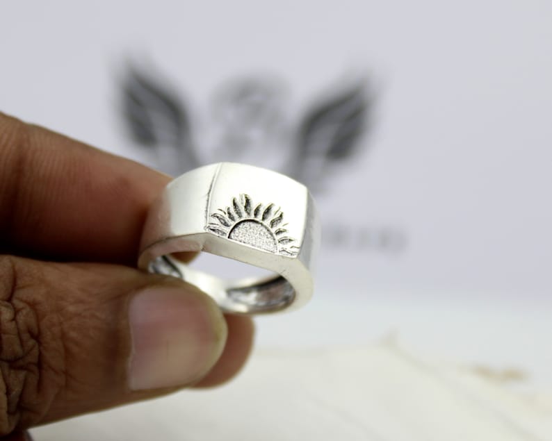 May include: A silver ring with a square-shaped band and a sun design engraved on the top.