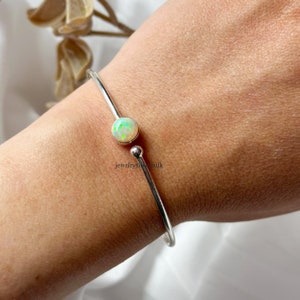 Może przedstawiać: Srebrna bransoletka typu bangle z jednym białym opalem. Bransoletka jest otwarta i ma prosty design.