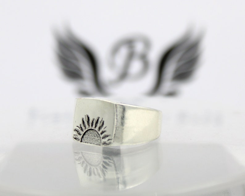 May include: A silver ring with a square face featuring a raised, detailed design of a sun rising over a horizon.