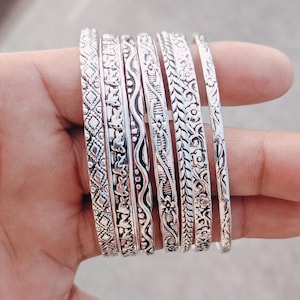 Könnte beinhalten: Eine Sammlung silberner Armreifen mit aufwendigen, geprägten Designs. Die Armreifen weisen verschiedene Muster auf, darunter florale, geometrische und wellenförmige Linien. Die Armbänder werden zusammengehalten, was die detaillierte Handwerkskunst und die reflektierende Qualität des Metalls zeigt.