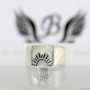 May include: A silver ring with a raised design of a sun on the top. The ring is wide and has a square shape.