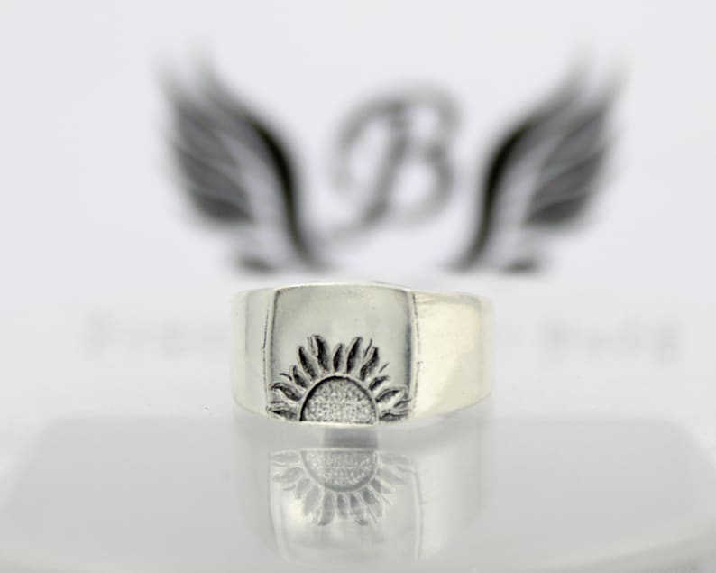 May include: A silver ring with a raised design of a sun on the top. The ring is wide and has a square shape.