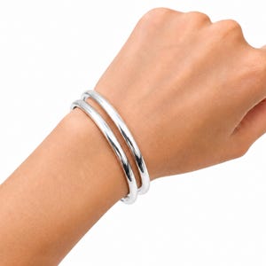 May include: A pair of polished silver-tone bangle bracelets with a smooth, rounded profile. The two identical circular bands feature a high-shine reflective finish and a simple, continuous design that sits comfortably around the wrist area.